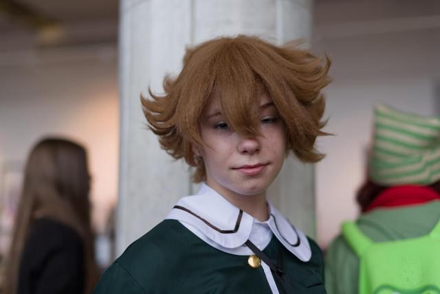 The image features an individual with reddish-brown hair styled upwards, standing indoors at a gathering that appears to be related to cosplay or costume events. The person's expression is contemplative and slightly downward-focused. They are dressed in a detailed green garment resembling a school uniform from a fictional setting, complete with white collars accented by brown trimmings and a gold-colored circular button on the chest.

The background features blurred figures: one with long brown hair in a black top, and another wearing a green-and-white striped hat, a red scarf, and a green vest. The setting appears to be an indoor space with a light-colored column visible. The main subject is in sharp focus, while background elements are out of focus.