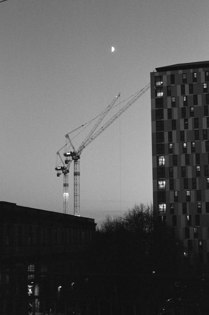 Half moon above a crane that's next to an apartment building and above another building.