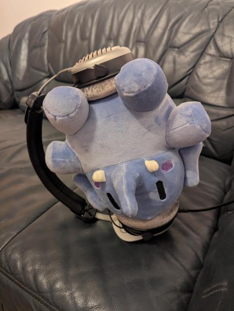 Small Blue Plush Mastodon, upside-down on the couch, with one headphone earpiece on her head, the other on her butt