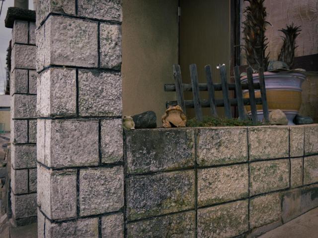 The image shows a porch: a stone structure with a low wall and adjacent pillar, both made of large, rectangular, light gray stones with dark mortar lines. The wall has a slightly weathered appearance with some discoloration. On top of the wall, there are small rounded objects (some rocks and two small clay frog sculptures) and sparse greenery. Behind the wall, a wooden picket fence with vertical slats is visible. To the right of the fence, large ceramic plant pots are present; one has a yellow and blue striped design. The background includes a beige wall and a door. The door is opened slightly, but interior is not visible.