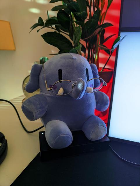 A blue mastodon plush sitting on a USB hub in front of a zamioculcas plant and wearing glasses.