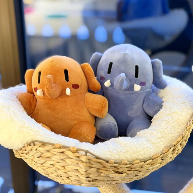 Two plushtodons, one orange and one blue, are sitting together in a soft, white, fluffy cat-tree  basket. Both have rounded bodies, small arms and legs, black vertical eyes, blushed cheeks, and little white tusks.They are V smol and incredibly cute!