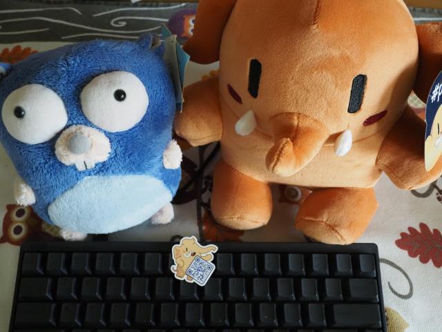 An activity peach mini Mastodon is pausing with a blue Go Gopher and a joinmastodon sticker behind an HHKB.
