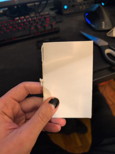 photo of a DIY pamphlet made from simple paper bound by thread. it's in someone's hands whose fingernails are painted black, albeit quite chipped. in the background is a  computer desk and some peripherals thereof