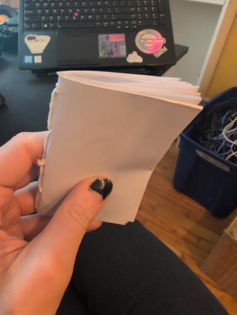a photo of a DIY pamphlet, or notebook, in someone's hand. it's made of simple paper and bound by thread, and is an eighth the size of standard printer paper

the person's hand's thumb has chipped black nail polish. in the background is a computer desk, a laptop with some stickers (a bowl of ramen, a scene from SLC punk, and the laughing man from ghost in the shell), as well as a large blue bin filled with cables