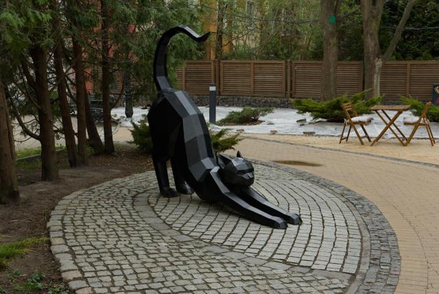 The image captures a large, geometrically designed statue of a cat in an outdoor setting. The sculpture is primarily black with gray accents and appears to be made from multiple overlapping triangular panels that give it a faceted appearance reminiscent of paper or cardboard cutouts. The cat is depicted mid-stretch on one side, as if caught during play or relaxation.

The surrounding area includes a paved circular path around the base of the statue, indicating this might be in a park or garden. In the background, there's an outdoor seating arrangement with wooden chairs and tables, suggesting a public space where people can sit and enjoy their surroundings. The environment looks calm, with bare trees and fallen leaves scattered on the ground.

There is no direct sunlight visible; it seems overcast which might explain why this area appears tranquil despite being in what could be an urban setting.