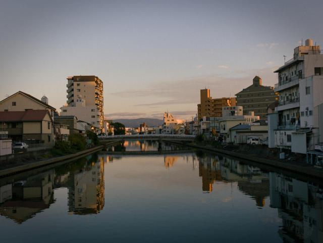 A photo taken from a bridge. The image shows a calm river running through an urban area. The water is still, creating clear reflections of the surrounding structures and sky. On both sides of the canal, there are multiple buildings: low-rise residential houses on the left, and a mix of mid-rise and high-rise residential/commercial buildings on the right. A bridge spans the canal in the middle distance. The sky displays a soft gradient of blue and light orange near the horizon, indicating twilight. Faint outlines of mountains or hills are visible in the distant background. The reflection in the canal mirrors the buildings and the sky accurately.