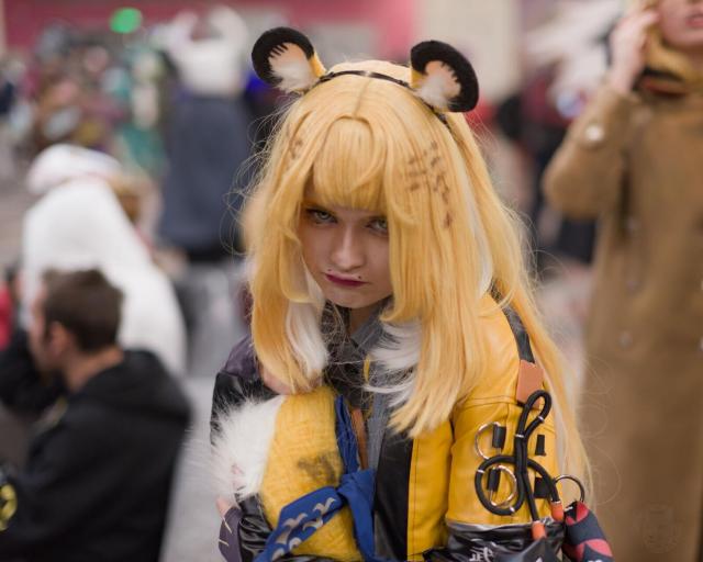 The image features a person in a costume as the central focus. They have long, blonde hair with white tips and bangs, and small brown markings are visible on the hair. Two rounded furry ear accessories (black on top, light brown and white on the inside) are attached to a headband. They wear a yellow jacket with black stripes and leather-like sections. The background is blurred, showing other people in various outfits (e.g., white, black, and brown coats), indicating a busy public setting, likely a convention or event.