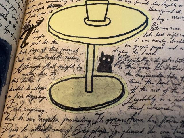 a sticker of a vase on a table, a drawing of a cat, and messy handwriting all around it featuring a failed attempt at a drop cap when I tried adopting a slightly different style of writing (more prominent capital letters, something like Spencerian script)