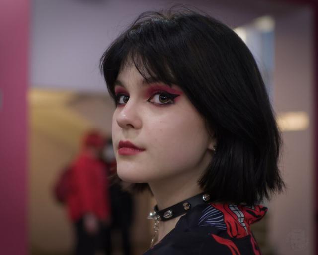 The image captures a young woman in profile, her gaze directed off to the side. She has short black hair and is wearing bold makeup with red eyeshadow that matches her vibrant lipstick. Her outfit includes a choker adorned with silver studs around her neck and she's dressed in a blouse featuring an intricate design on one shoulder.

The background suggests an indoor setting, possibly a public space like a museum or gallery, indicated by the blurred figures of people moving about behind her. The overall mood is contemplative and artistic, enhanced by the contrast between the clarity with which the woman is portrayed against the soft focus of the backdrop.
