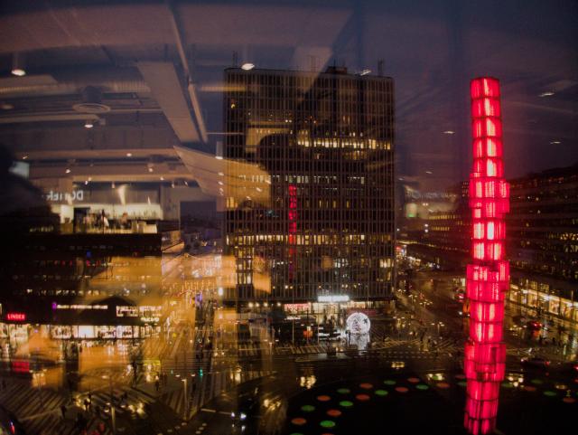 Sergels torg (Sergel square) in central Stockholm, seen through a window with reflections from behind. On the left side is the reflections is dominant, on the right side a read sculpture is dominant. In the background tall office buildings.