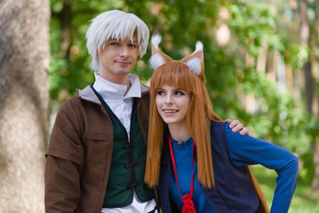 The image captures two individuals in what appears to be a cosplay scenario. The person on the left is dressed in attire reminiscent of medieval West European clothing. This includes a brown and white garment layered over a green vest and white shirt.

The individual on the right sports features typical of an animal-inspired character costume; specifically resembling fox traits with red fur and pointed ears. They are dressed in what appears dark-blue vest and a vibrant blue top with a collar and long sleeves. The background suggests they are outdoors during daylight, possibly in a park or forested area given the greenery visible.