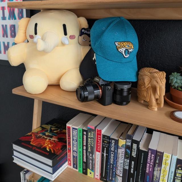 A photo of a bookcase with assorted books and items, focused on a yellow Mastodon Plushie. The books are mostly sci-fi, and include some of my favorites, such as The City We Became by N. K. Jemisin, The Long Way to a Small, Angry Planet by Becky Chambers, and Mr. Penumbra's 24 Hour Bookstore by Robin Sloan.