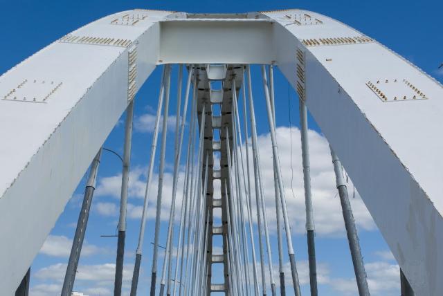 The image captures a close-up perspective of a white suspension bridge, focusing on the vertical steel cables that are intricately arranged in a symmetrical pattern. These cables converge at two points - one towards each side where they connect with large truss sections and end caps painted in shades of gray and red. The sky above is clear blue with minimal cloud cover, providing a contrasting backdrop to the metallic sheen of the bridge's structure.