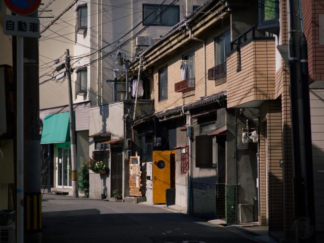 This image depicts a narrow urban alleyway in a Japanese city. On the left, a vertical sign with Japanese text is visible, mounted on a pole near overhead wires. A building with a green awning and potted plants sits further left. On the right, multi-story residential buildings feature brick and concrete facades; one has a yellow door, balconies with hanging laundry, and windows with air conditioning units. Small awnings, a wooden structure near the yellow door, and green tiles on part of a building are also present. The paved street is empty, illuminated by daytime light with visible shadows.