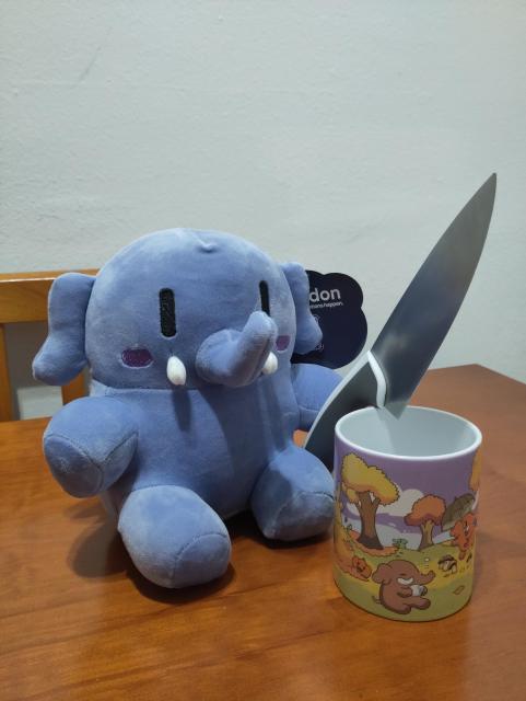 Fotografía del peluche azul de Mastodon, junto a la taza con decorados otoñales de Mastodon. El peluche parece que está sosteniendo un cuchillo más grande que el propio peluche.