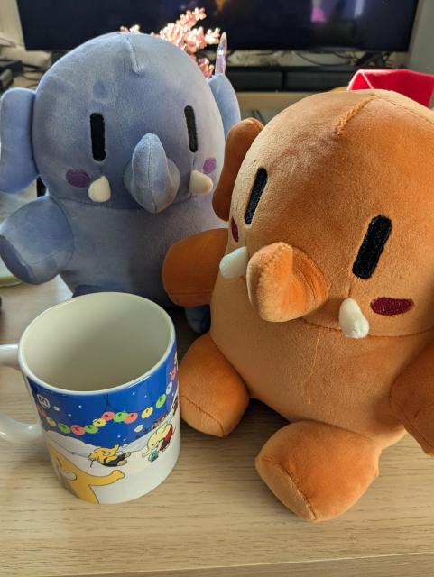 the purple and orange mastodon plushies sitting on a table. There's a mastodon coffee cup next to them.