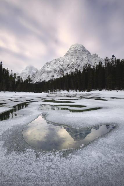 A snowy winter scene taken ontop thin ice. Holes in the ice reflect the sunrise light and the snowy peak in the distance. The ice over the river is covered in thin ice with frost ontop.