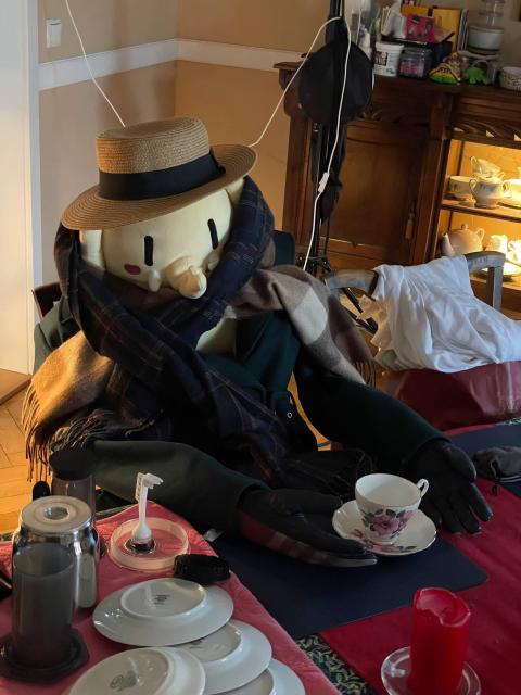 The #Plushtodon XL sitting at the dinner table while wearing a coat, a scarf, a hat, and gloves, resembling a person, with an empty tea cup in front.