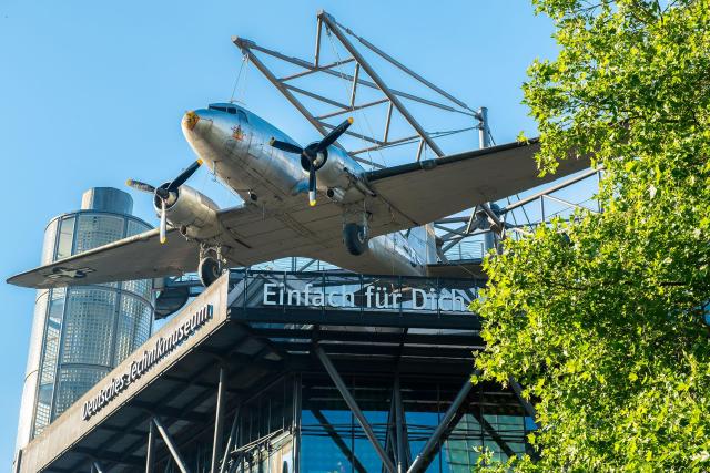 Ein silbernes Flugzeug hängt an einer Metallkonstruktion an einer Gebäudefassade. Darunter befindet sich eine Aussichtsplattform, an der am Geländer der Schriftzug "Deutsches Technikmuseum" und "Einfach für Dich" angebracht ist. Auf der rechten Seite sind grüne Zweige, im Hintergrund blauer Himmel.