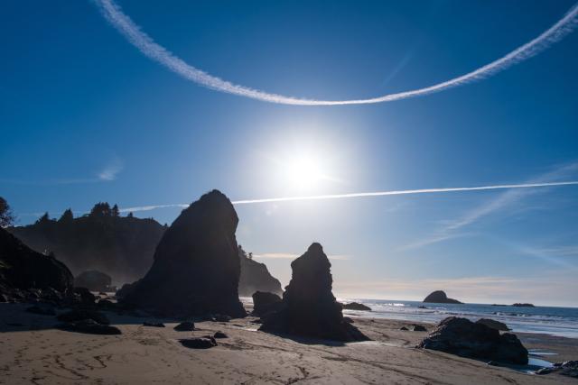 Big rocky formations on the beach at sunset, with a circular contrail overhead. [Fuji X-T5 / Fuji 35 1.4]