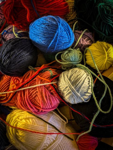 A vibrant, close-up shot of several balls of yarn in various colors and sizes, haphazardly piled together. 

The collection features a wide range of colors, red, blue, yellow, green, black and white.

The lighting is coming from the side, which creates deep shadows and highlights the fuzzy, twisted texture of the individual strands.