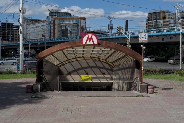 The image captures a scene from an urban setting, specifically highlighting the entrance to a subway station. The most prominent feature is the curved metal structure of the shelter. This metallic construction has a reddish-brown hue and sports a large "M" logo at its peak, signifying it as part of a metro system.

In the background, beyond the shelter and station entrance, one can see tall buildings reaching up into the sky. These structures contribute to a sense of depth and scale in the image. A blue overpass bridge also spans across this scene, hinting at an intricate network of transportation infrastructure within reach.

The sky overhead is partly cloudy but allows enough sunlight to brighten the overall ambiance, casting shadows on the ground below. The colors present are primarily urban tones - reds from the shelter and paving stones, browns and greys from the metallic construction, blues in the distance from the bridge and possibly some of the buildings.
