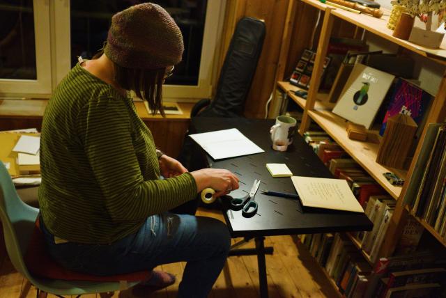 another person, also with a beanie, is sitting at a desk and doing something with scissors and stuff. 