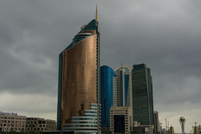 The image captures a dynamic urban landscape under an overcast sky. Dominating the scene is a tall, modern skyscraper with a unique design; its facade combines blue glass and golden hues that shimmer against the cloudy backdrop. The building's distinct architecture suggests it could be a landmark or significant structure within this cityscape.

In the vicinity of this central skyscraper are other contemporary high-rise buildings, featuring sleek designs typical of urban development in major cities. Their presence adds to the sense of progression and modernity captured by the image.

The overcast sky casts a soft light on the scene, muting colors slightly but enhancing the textures of the building materials. The cloud cover also hints at possible weather changes, adding an element of anticipation or drama to the setting.

Overall, this urban snapshot paints a picture of a bustling city with significant architectural achievements and a dynamic atmosphere under changing skies.