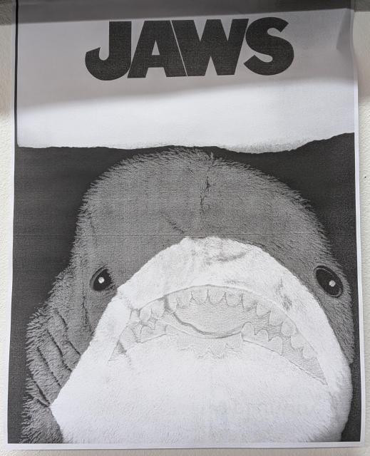A b/w photocopy image of a small blahaj with the text "JAWS" above, printed on a piece of paper with a torn edge. The blahaj is squished against the glass of the copier. The composition is reminiscent of the movie poster.
