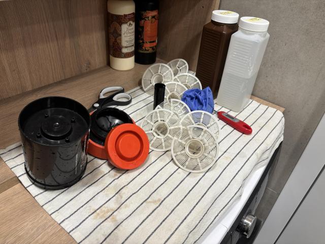 A workspace with various items is displayed, including a black and red container, several white fan parts, scissors, two bottles, a latex glove, and a thermometer, all arranged on a striped towel. A wooden shelf in the background holds decorative bottles.