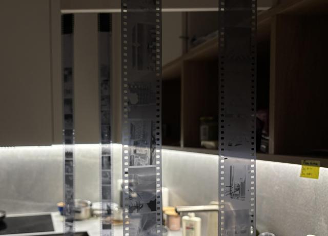 The image shows black and white film strips hanging in a dimly lit kitchen. The background features shelves with various kitchen items and a soft light illuminating the countertop area.
