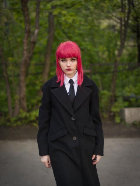 The picture features a young woman standing outdoors in a natural setting, possibly a park or a garden. She is wearing a black coat with  two front pockets. Underneath the coat, she's wearing a white shirt with a black tie. The woman has vibrant pink hair styled in a straight, shoulder-length cut. She is facing the camera directly, giving an engaging portrait-like quality to her appearance. The expression on her face is serious, which may suggest an underlying narrative or emotion related to the context in which this photograph was taken. The background consists of green foliage and trees, suggesting a serene and natural environment. The overall atmosphere of the image is calm and somewhat contemplative.