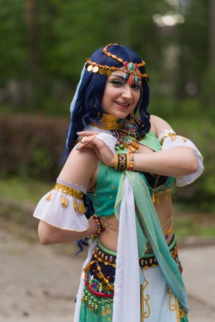 The image features a woman dressed in heavily stylised Arabic attire, standing against the backdrop of a park. She is dressed in an elaborate and ornate costume, featuring a lavish headdress. The costume is richly detailed with gold trimmings, colorful gemstones, and sequins adding sparkle to her ensemble. Her hair is styled into long waves, dyed in a striking shade of blue, which contrasts vividly against the lush green backdrop of trees seen through what appears to be an outdoor setting. The woman's face is lit up with a smile as she poses for the camera.