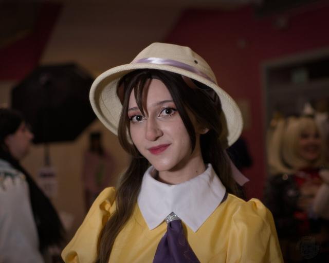 The image captures a young woman standing against the backdrop of what appears to be an indoor event, possibly a convention or gathering. She is dressed in a costume reminiscent of anime or manga culture; her outfit includes a yellow dress with purple accents and a white collar adorned with a sparkling brooch at its center. Adding character to her ensemble is a beige bucket hat decorated with a lavender ribbon around the crown.

The woman's expression is one of mild amusement, as she looks directly into the camera lens. Her hair is styled in loose waves that cascade over her shoulders, and she wears makeup that accentuates her features without overpowering them. The lighting in the room casts soft shadows on her face, adding depth to her appearance.

In terms of background context, although blurred, it suggests a social setting with other people present. Some individuals can be seen wearing costumes or props typical of cosplay events.