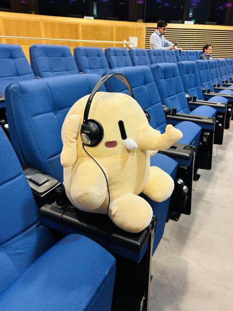 The mastodon plushie (a little elephant) sits on a blue chair in a row of blue chairs, wearing a translation headset 