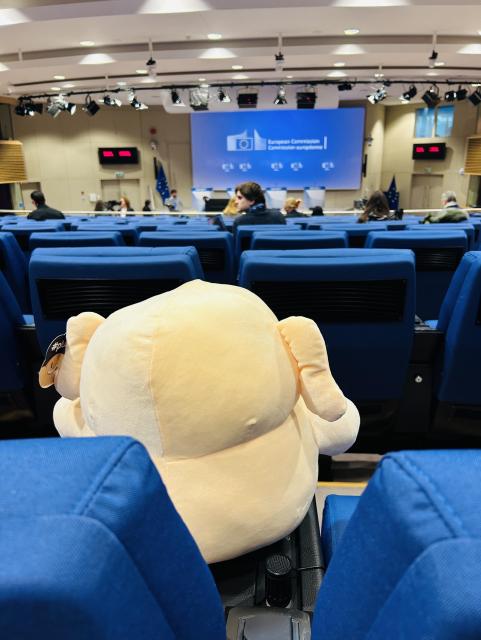 Mastodon plushie photographed from behind, hes sitting in the press room, in front of him are a couple of journalists and a screen featuring the European Commission logo and some podiums 