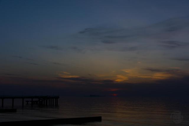 The image depicts a serene sunset over the sea. The sky is painted in shades of blue, with wispy clouds scattered across it. The sun is setting behind the horizon, casting a warm glow on the water below. In the foreground, there's a pier extending into the distance.

The calm waters reflect the vibrant colors of the sky and the sun, creating a beautiful mirror image effect. The perspective of the photo is taken from the shoreline, looking out towards the sea, giving a sense of depth and vastness to the scene.
