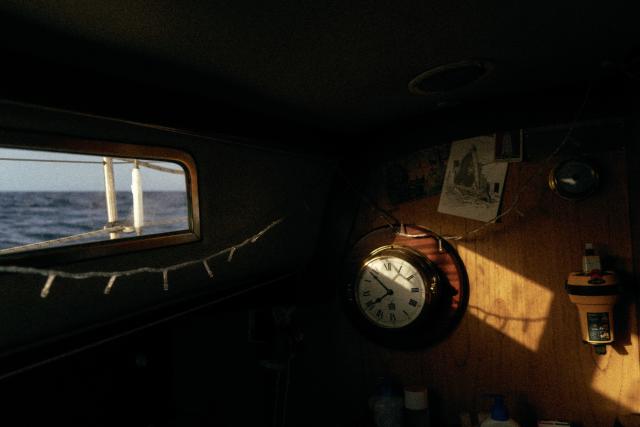 The interior of a boat at sea: through the portlight, an empty sea; a ray of light illuminates a wooden bulkheadm with a large ship's clock and an emergency beacon.