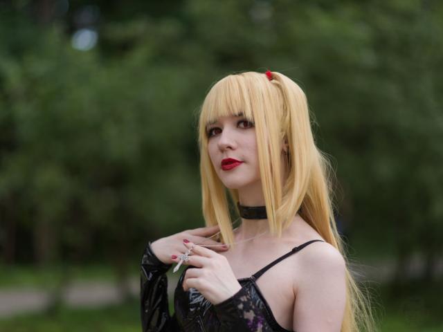 The image features a young woman with blonde hair, dressed in black attire. Her makeup is done dramatically, featuring bold eyeshadow and lipstick. She's wearing a black outfit with lace details, which gives it an elegant yet edgy look. A notable feature of this attire seems to be a choker around her neck and cross-shaped pendant she supporting by her left hand fingers. The woman's expression is curious as she gaze directly into the camera while touching her collarbone by her right hand.

She is standing against the backdrop of a park or garden setting, surrounded by trees that provide a natural and serene atmosphere to her appearance. The woman's pose suggests she might be posing for a photo.
