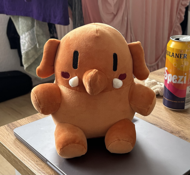 A Mastodon mini plush elephant colored "Activity Peach", a strong orange. It sits atop a MacBook Pro, to the side of a Spezi soda can, because yes.