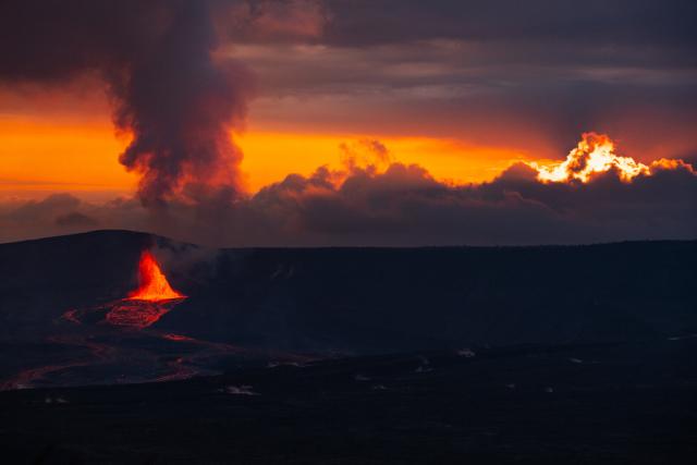 A volcano erupts amidst a dark rocky landscape, with a sunset obscured by clouds behind. [Fuji X-T5 / Tamron 18-300]