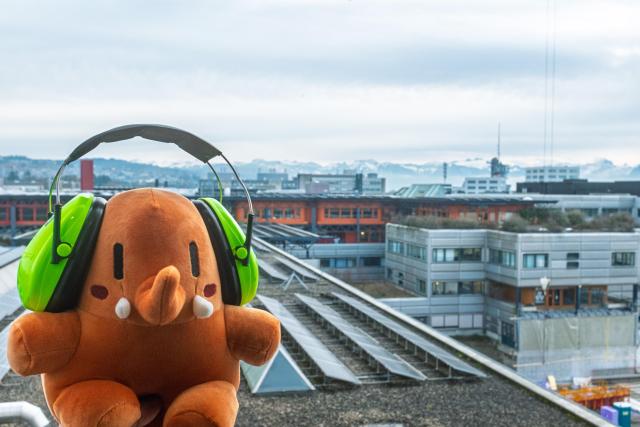 La mascotte Plushtodon, au-dessus des toits de l'EPFL, avec des casques anti-bruit sur les oreilles. Les Alpes en arrière-plan.