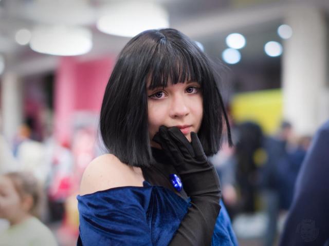 The image shows a young woman in a cosplay costume, likely at a convention or event. The scene is somewhat blurred, with a shallow depth of field that emphasizes the woman as the primary subject. 

The young woman has dark hair, styled in a short, choppy bob. Some strands fall forward, partially covering her face. Her skin tone appears pale, and she has dark, expressive eyes. She is looking directly at the camera with a slightly serious expression. She is wearing a dark blue velvet off-the-shoulder dress. A black glove is covering her left hand, and she is also wearing a large, dark blue, possibly crystal, pendant. Her head is turned slightly to the side, and she has a hand gently covering her mouth.

The background is blurred and dark, with indistinct shapes and colors. There are what appear to be circular lights, possibly from stage lighting or decorations, visible in the upper right corner. Some people can be seen in the blurred background.