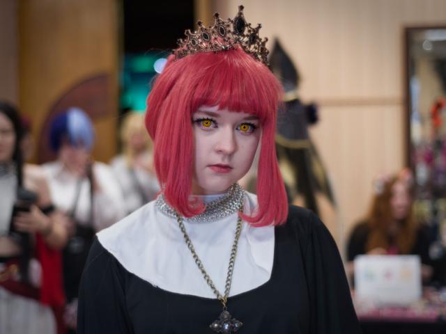 The image shows a young woman dressed as a character, likely from a popular anime or manga series. She has vibrant, bright red shoulder-length hair styled with a slight wave or curl. She is wearing dramatic makeup, including dark eyeshadow, and striking yellow contact lenses, which are a key element of the look. She's wearing a black dress, a white collar, and a silver chain necklace around her neck with a gold chain that features a large, ornate pendant. The woman is also wearing a tiara on her head. In the background, there are other people present but they are blurred out, drawing focus to the main subject in the foreground.