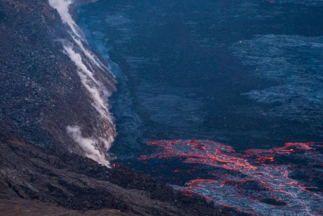 Flowing lava in a crater meets the wall, with steam emanating from the join. [Fuji X-T5 / Tamron 18-300]