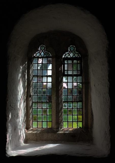 A church window with small panes in a deep recess. Some of the panes are clear and others lightly coloured. Sun is streaming through the window at an angle, lighting up the whitewashed wall and casting shadows of the window.