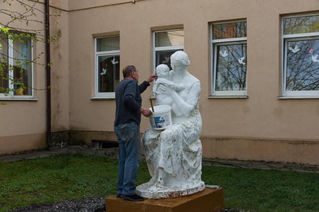 In the image, a man is meticulously painting a white statue of a mother and child. The statue depicts a woman holding her child in her arms, with both figures facing each other. The man stands to the left side of the statue, using a bucket filled with paint to add color to the scene. The statue is situated on a pedestal, which rests on a patch of grass. In the background, there are several windows that suggest that the scene is taking place in some sort of yard or patio.
