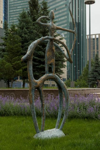 The image features a large, bronze statue of a man holding a bow in his left hand. The man is depicted as a very stylised figure with a long head and thin hands. He stands on the back of some stylised animal, like antelope, between its long curled horns, facing towards the right. All the sculpture resembles some kind of ancient petroglyph.

The statue is situated in an outdoor setting, surrounded by lush green grass and trees. In the background, there are tall buildings visible behind the man's figure.