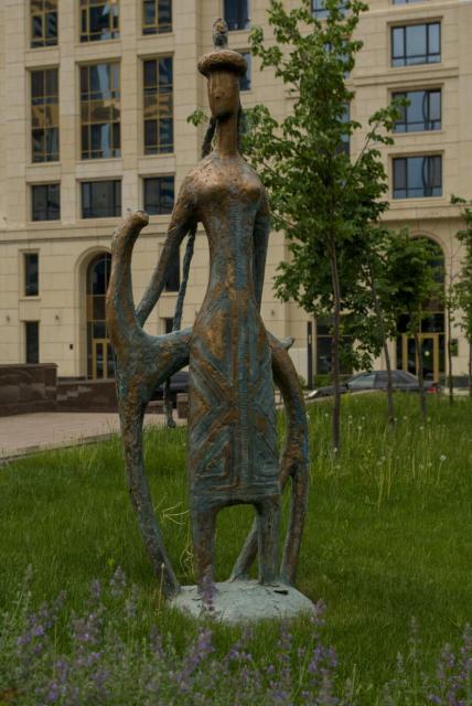 The image features a bronze statue of a stylised woman standing in a grass with her hands placed behind her on the neck and back of a thin and high dog standing behind her. All the sculpture resembles some kind of ancient petroglyph.

The statue is situated in a grassy area with purple flowers nearby. In the background, there are some young trees and a yellow building visible.